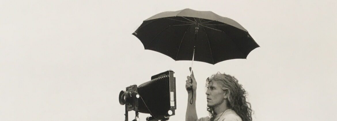 Der Fotograf Martin Blume steht auf einem Sockel an einem Kamerastativ und hält einen Schirm über sich. Schwarzweißfotografie.